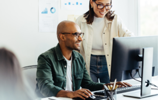 Two smiling business people stand over a desktop computer viewing the screen in an office, symbolizing Managed Network Services.