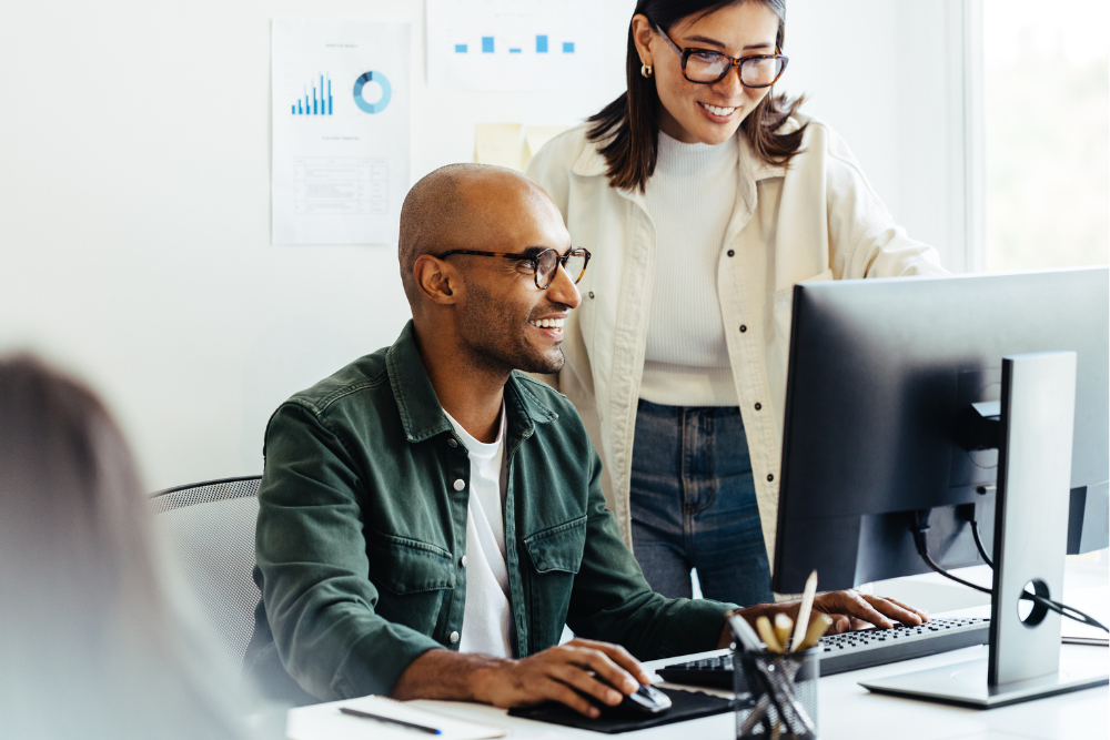 Two smiling business people stand over a desktop computer viewing the screen in an office, symbolizing Managed Network Services.
