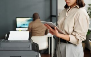 In an office environment, a woman is standing beside a multifunction printer, attentively using a tablet, indicative of the integration and monitoring aspects of Managed Printing Services that facilitate efficient workflow and document management.