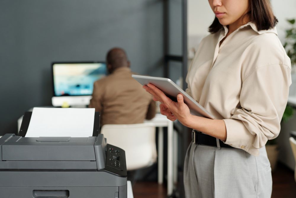 In an office environment, a woman is standing beside a multifunction printer, attentively using a tablet, indicative of the integration and monitoring aspects of Managed Printing Services that facilitate efficient workflow and document management.