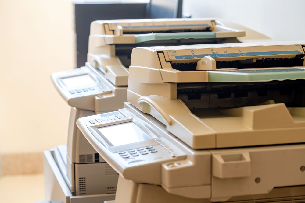 A close-up view of a line of high-end office copiers with document feeders and control panels, illustrating options for businesses considering a copier lease, against a soft beige wall.