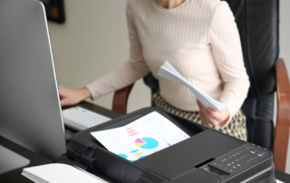 A woman working at her desk with documents and a colorful chart being printed from a nearby printer. This image illustrates the efficiency of Managed Print Services (MPS), showcasing how businesses can streamline their printing processes and manage documents effectively.