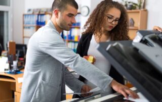 Two office workers using a multifunction copier, with one person operating the touchscreen and the other holding documents and an apple, highlighting the everyday use and convenience of a copier lease in a busy office environment.